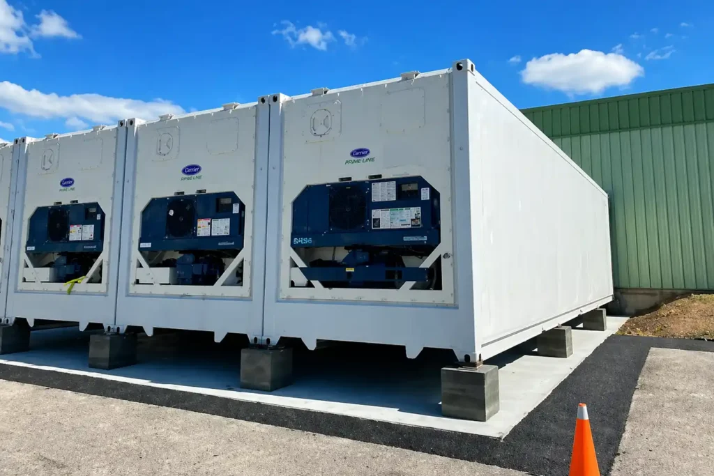 Multiple black shipping container levelling pads stacked to support the bottom corner of a sea can on uneven ground.