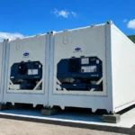 Multiple black shipping container levelling pads stacked to support the bottom corner of a sea can on uneven ground.